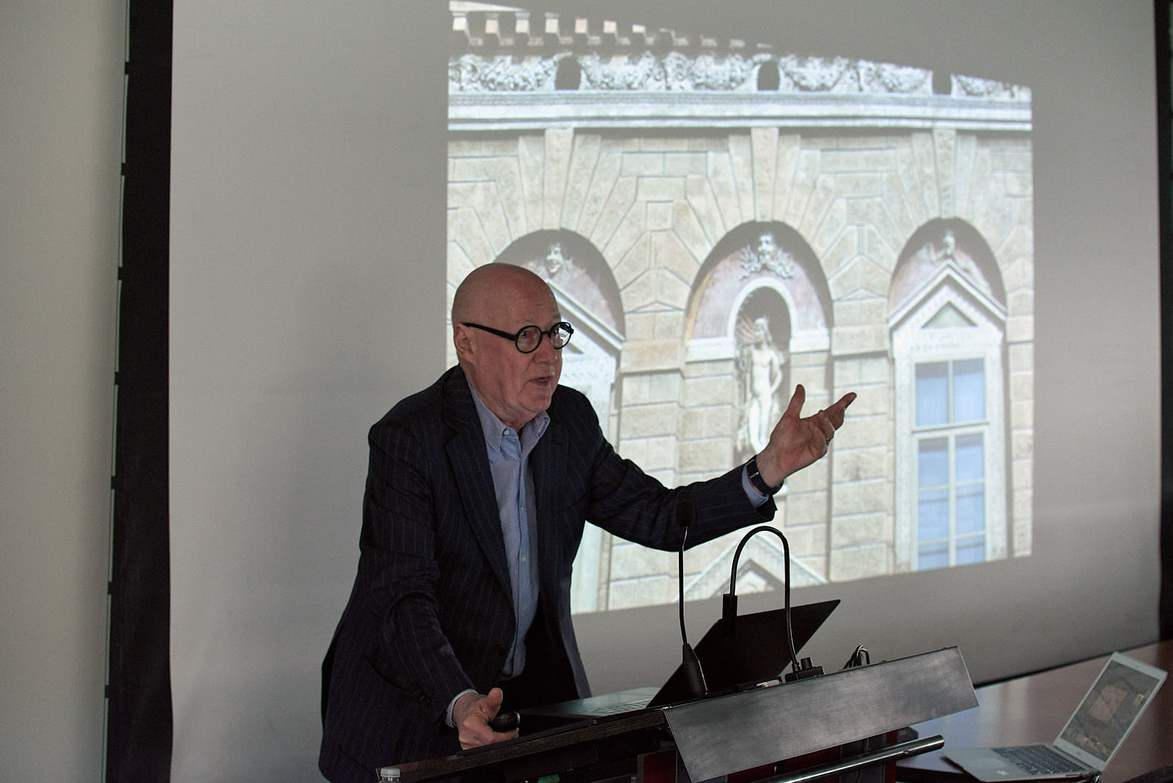 Man in jacket and glasses lectures in front of architecture slides